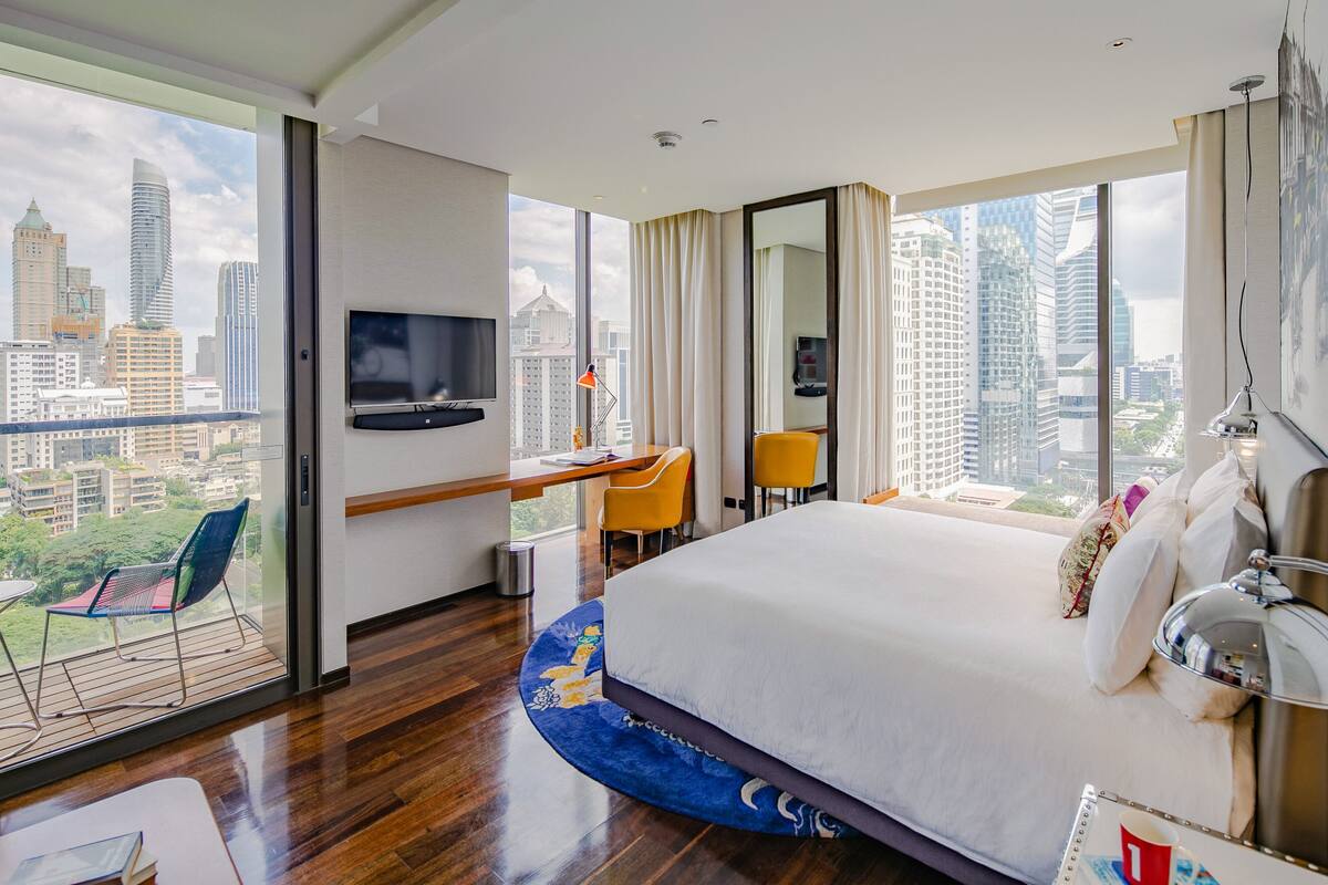 A hotel room with a large bed, a desk with a chair, a TV, and a balcony with a view of the city.