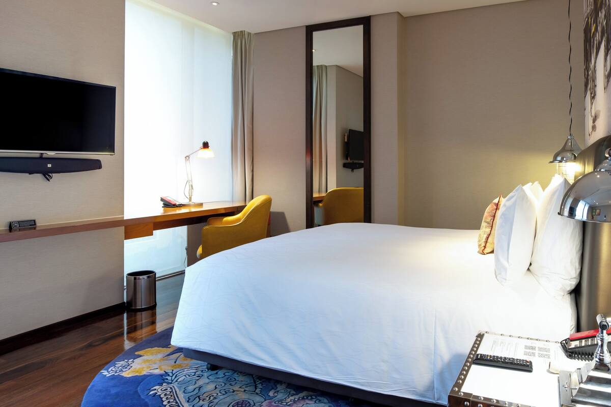 A hotel room with a bed, a desk with a chair, a television, a lamp, and a trash can.