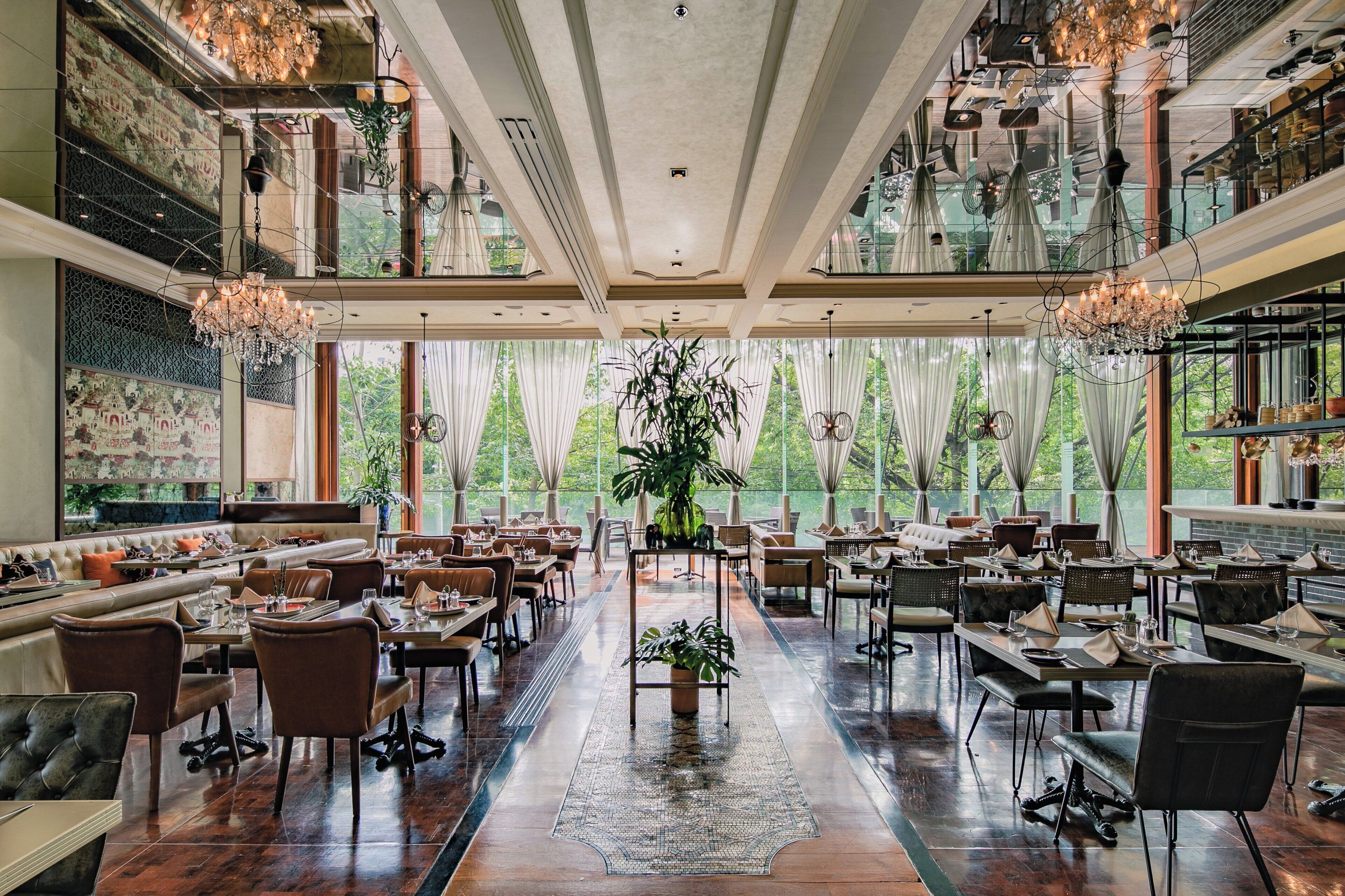 A spacious restaurant with high ceilings, chandeliers, and large windows offering a view of greenery.