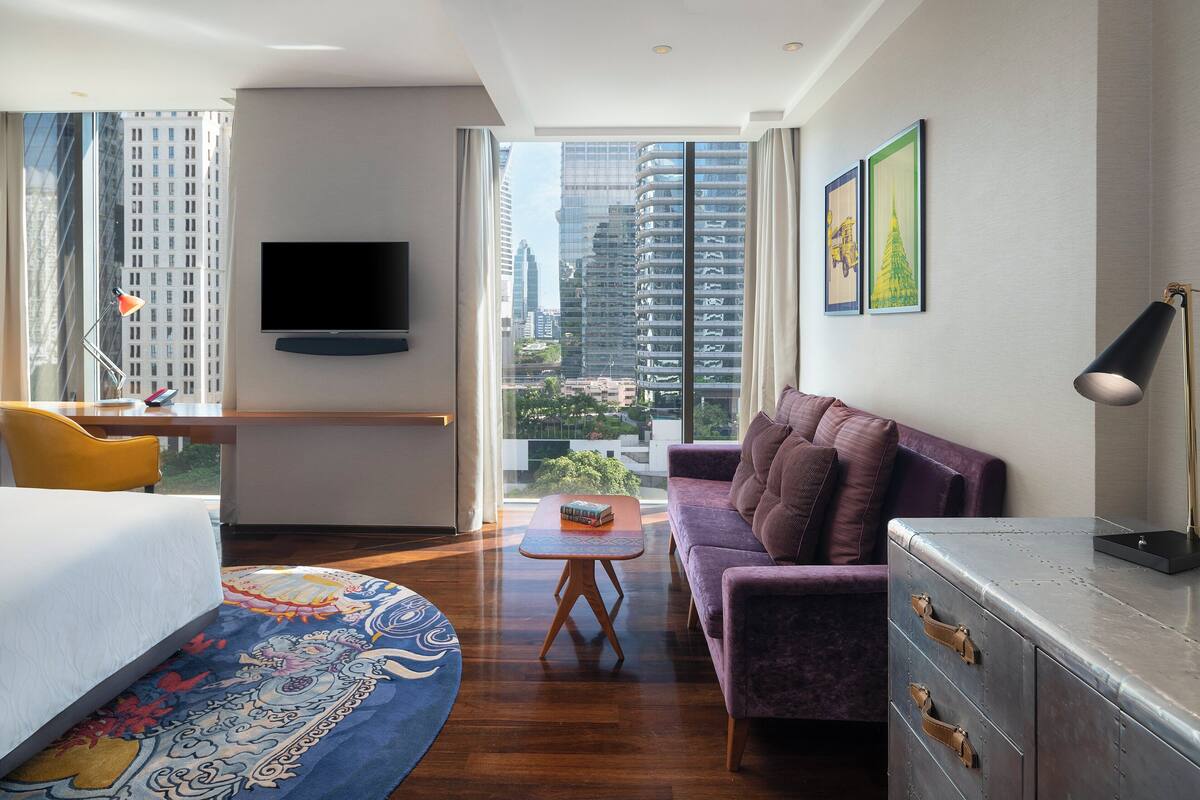 A modern hotel room with a large bed, a sofa, a small table, and a TV mounted on the wall.