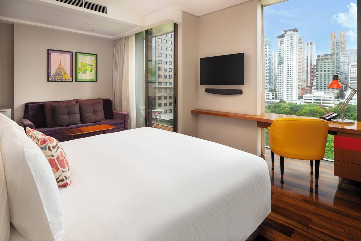 A hotel room with a large bed, a sofa, a desk with a chair, a TV mounted on the wall, and a window with a city view.