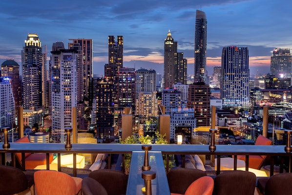 Vista panorámica de la ciudad desde una terraza en la azotea, con edificios modernos, una zona de comedor con sillas y mesas, y una barandilla.