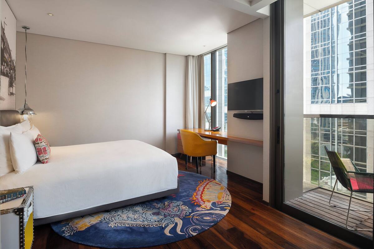 A modern hotel room with a large bed, a desk, a chair, and a balcony with a view of the city.