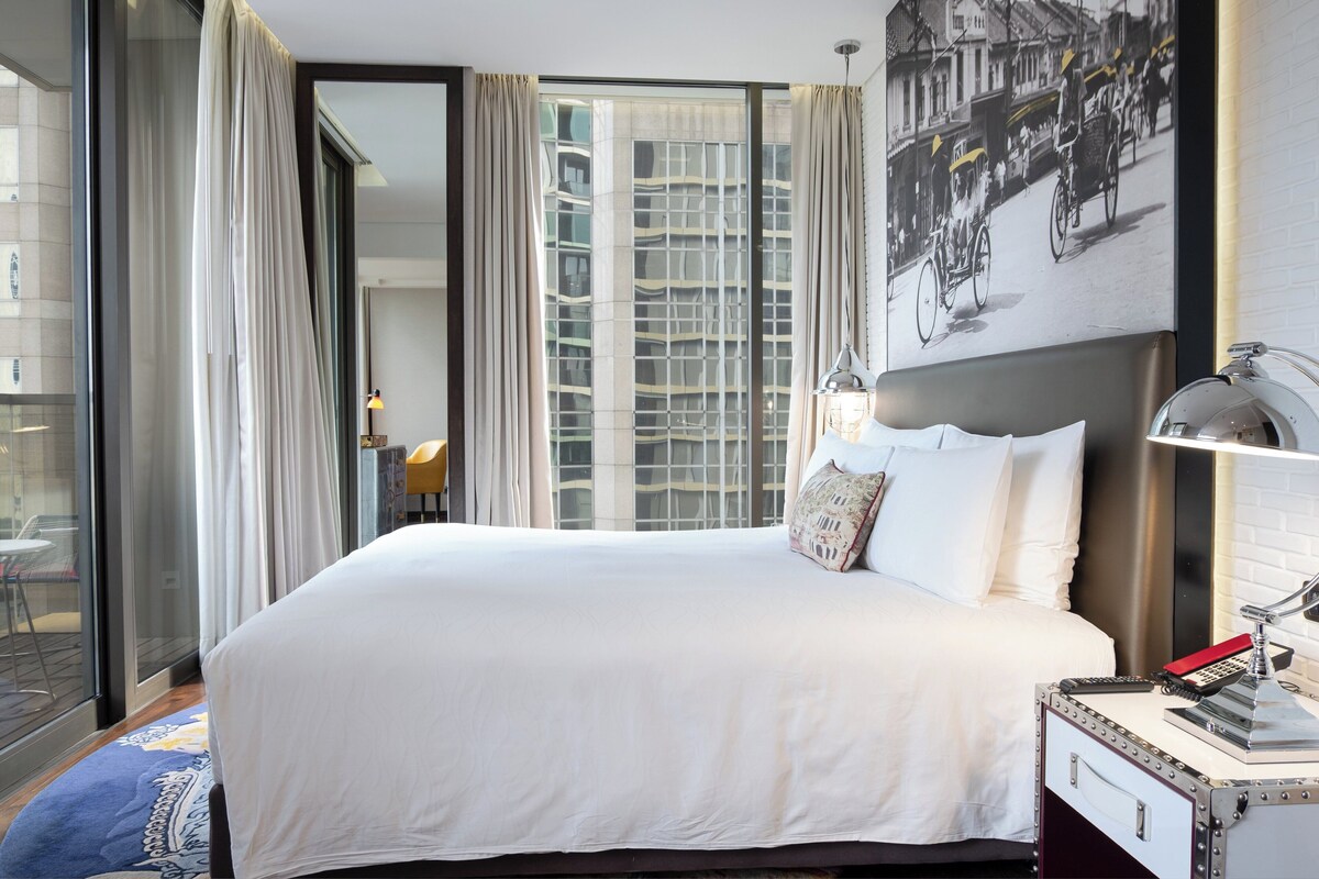 A hotel room with a large bed, a nightstand with a lamp, a window with curtains, and a wall-mounted picture.
