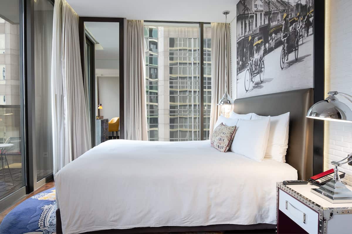 A hotel room with a large bed, a nightstand with a lamp, a window with curtains, and a wall-mounted picture.