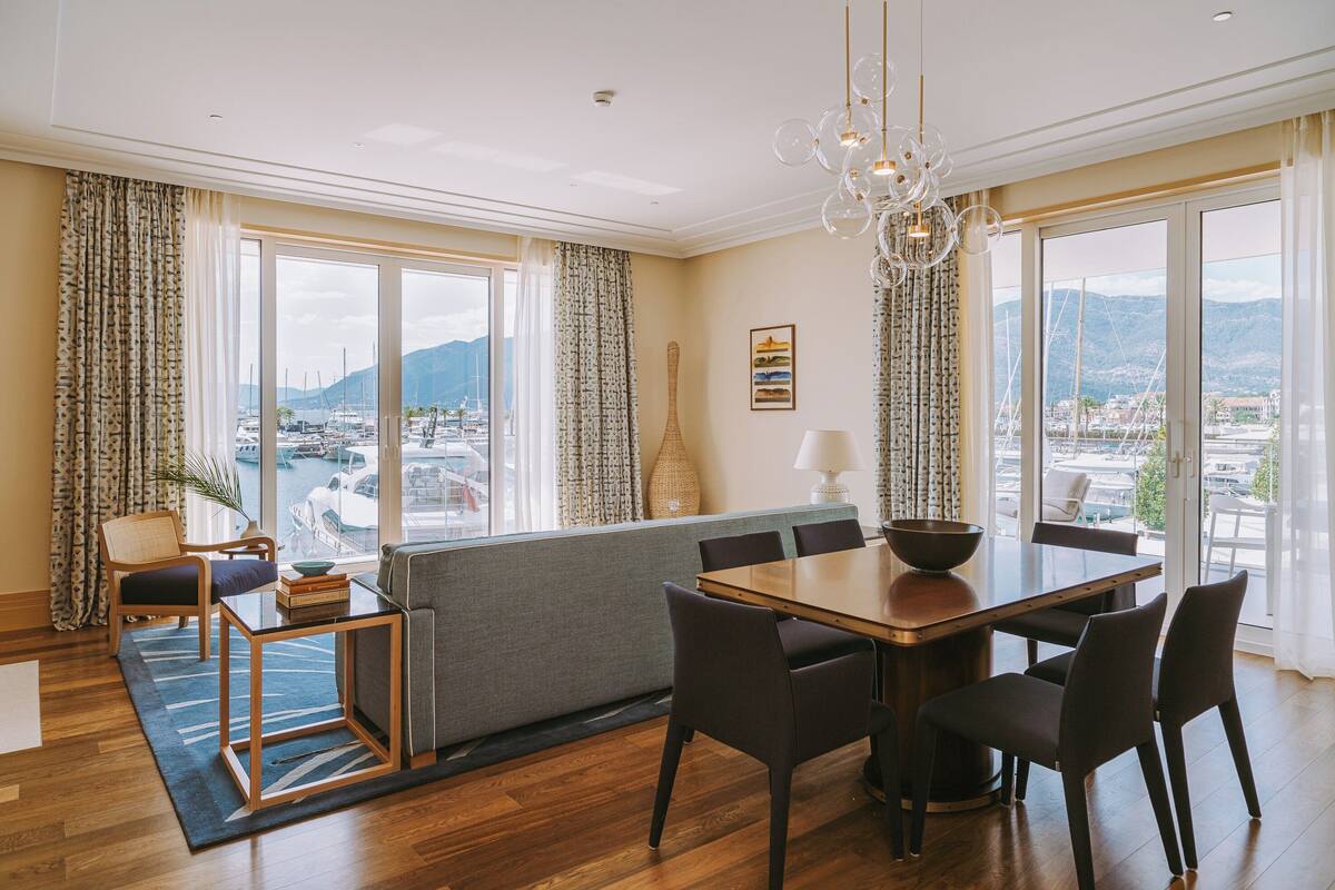 A modern dining area with a wooden table and chairs, a sofa, and a view of the marina through large windows.