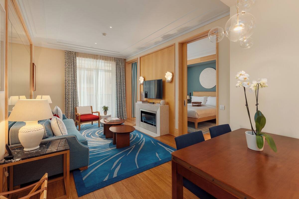 A modern hotel room with a sofa, a dining table, a TV, and a fireplace.
