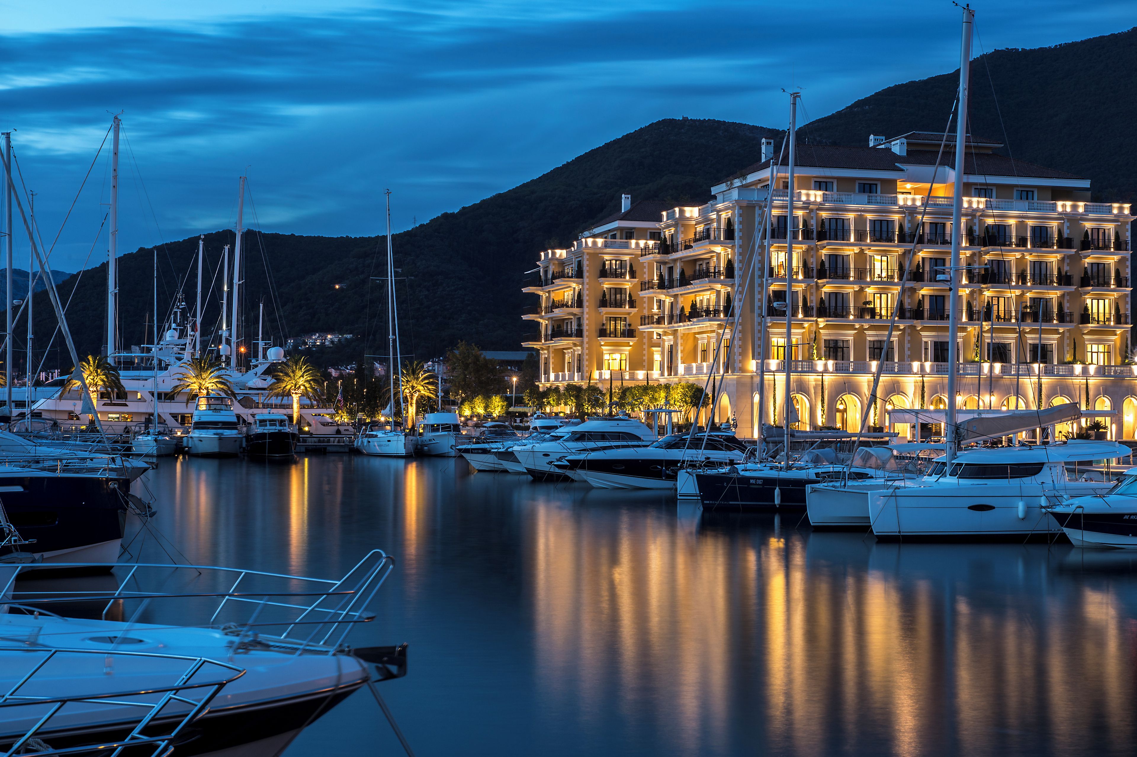 A marina with multiple yachts docked, a large illuminated building with balconies, and a mountainous backdrop.