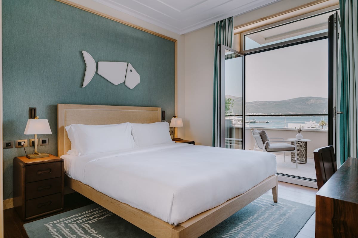 A hotel room with a bed, two bedside lamps, a nightstand, a desk, and a chair. There is a balcony with a view of the sea and mountains.