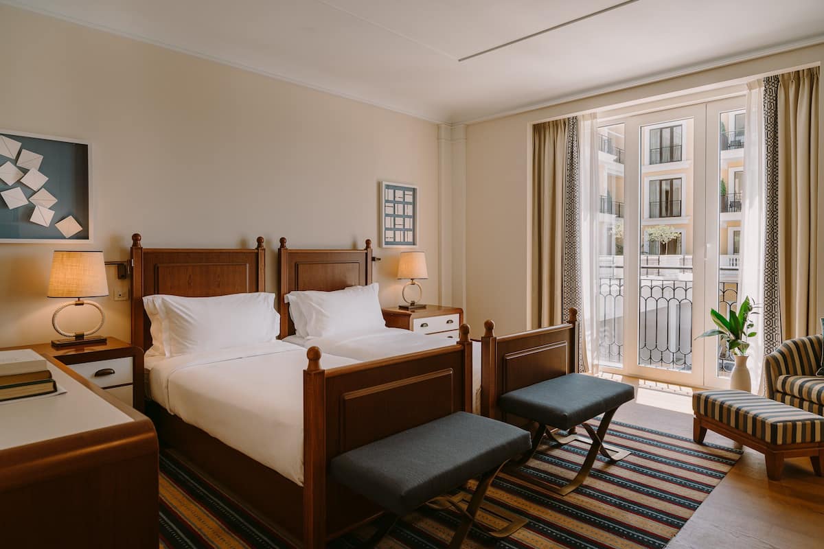 A hotel room with a wooden bed, bedside tables, a chair, a bench, a view of the outside balcony, and a window with curtains.