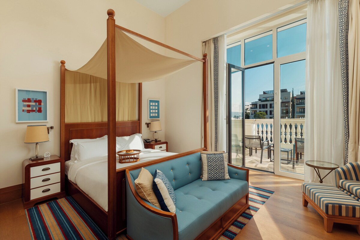 A four-poster bed with a canopy, a sofa, a chair, and a balcony with outdoor seating.