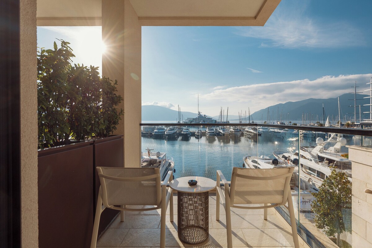 A balcony with two wicker chairs and a small table, overlooking a marina with boats and mountains in the background.
