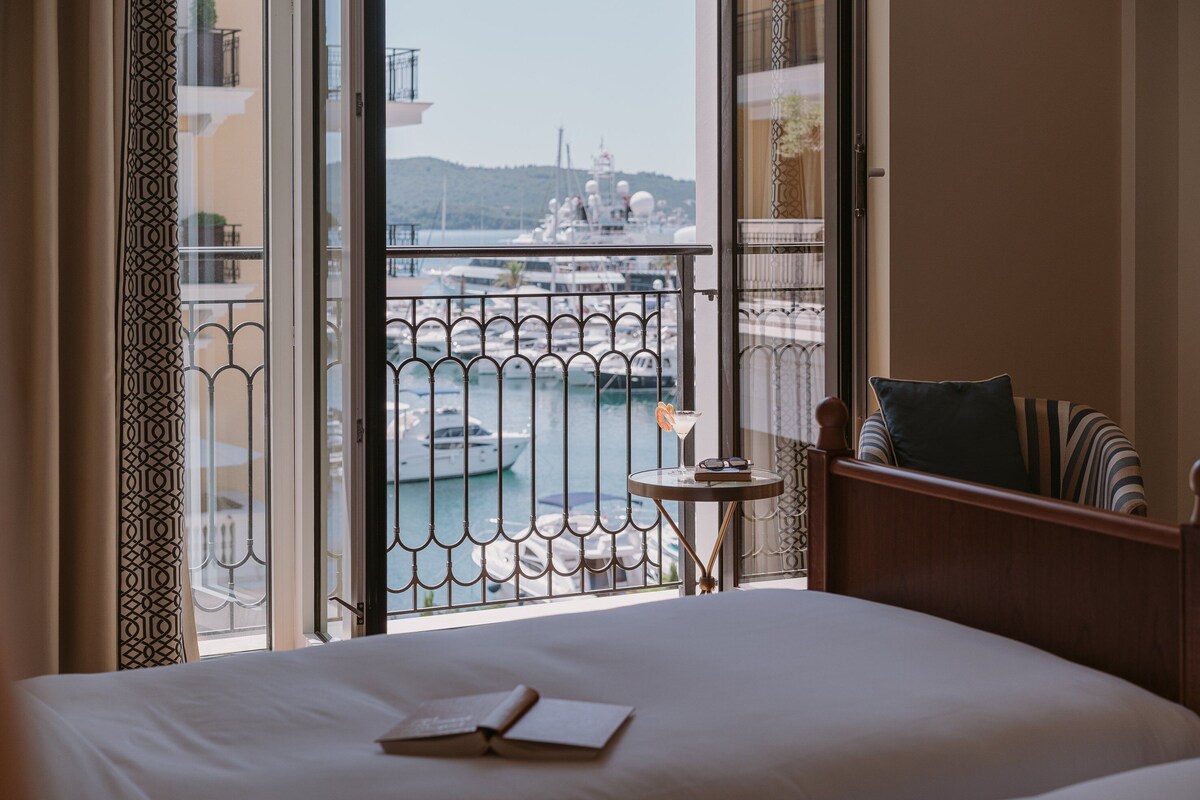 A hotel room with a large bed, a small table with a martini glass, and a view of a marina with boats.