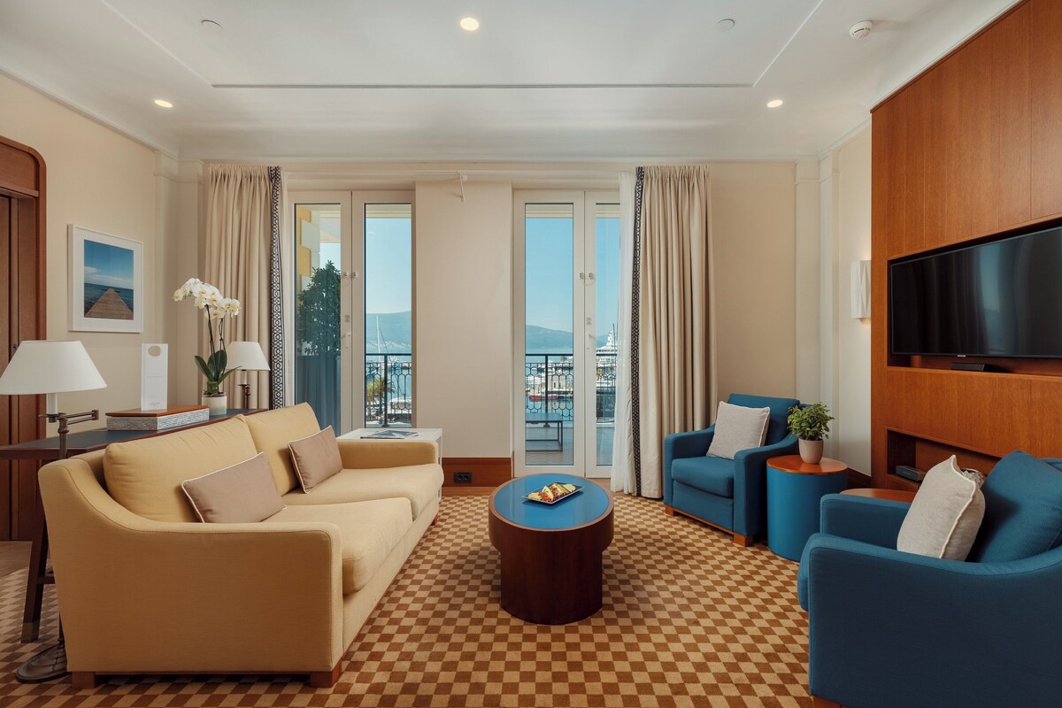 A hotel room with a checkered floor, a sofa, a blue armchair, a coffee table, a TV, and a balcony with a view.