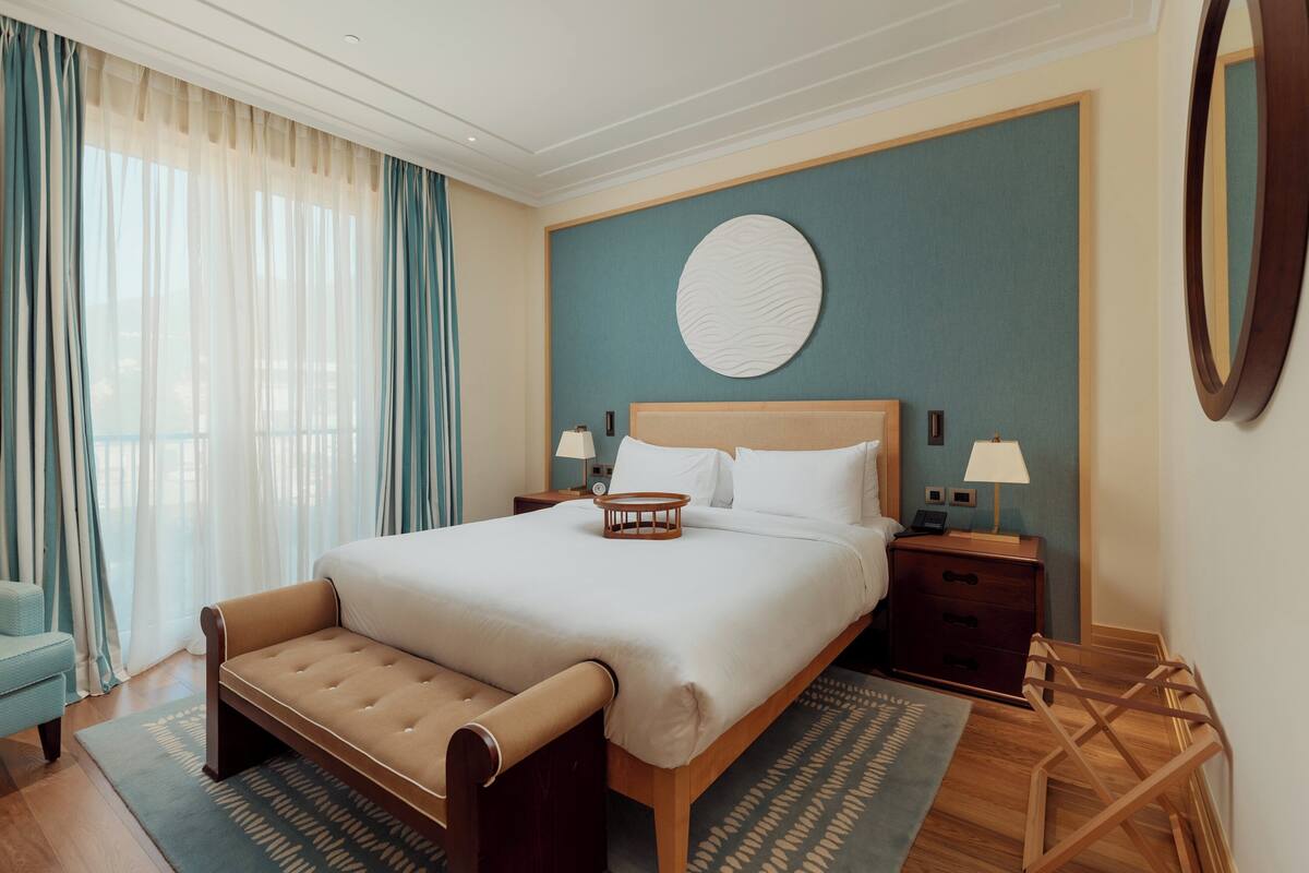 A neatly arranged hotel room with a large bed, bedside tables, a bench, and a view of the outdoors through sheer curtains.