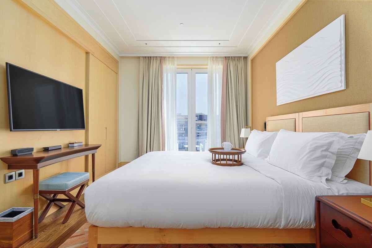 A hotel room with a large bed, a desk with a flat-screen TV, a small table with a cup, and a view of buildings through the window.