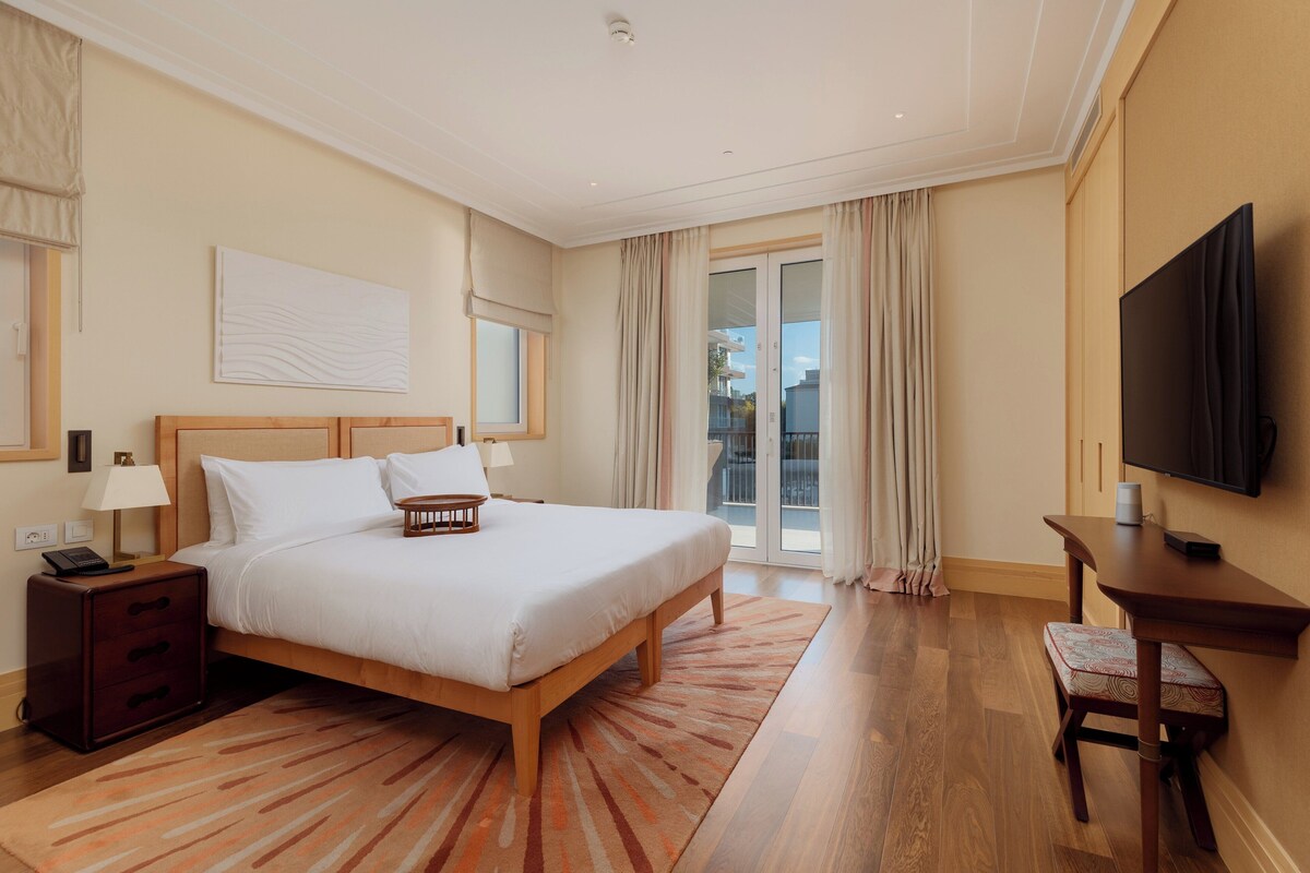 A hotel room with a large bed, a desk, a TV, and a balcony with curtains.