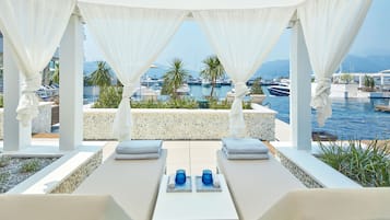 Un área de estar al aire libre techada con dos sillones, una mesita y vistas al puerto deportivo con barcos y palmeras.