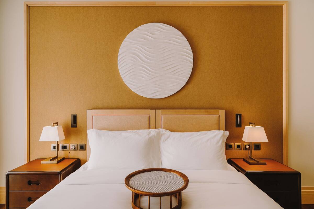 A hotel room with a bed, two bedside tables with lamps, a round rattan table, and a textured wall decoration.