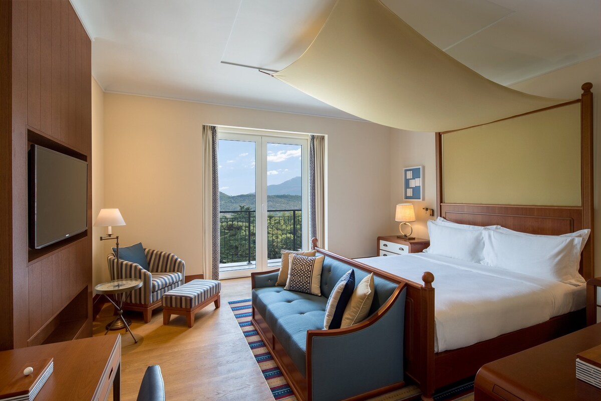A hotel room with a large bed, a sofa, a small table, a TV, and a balcony with a mountain view.