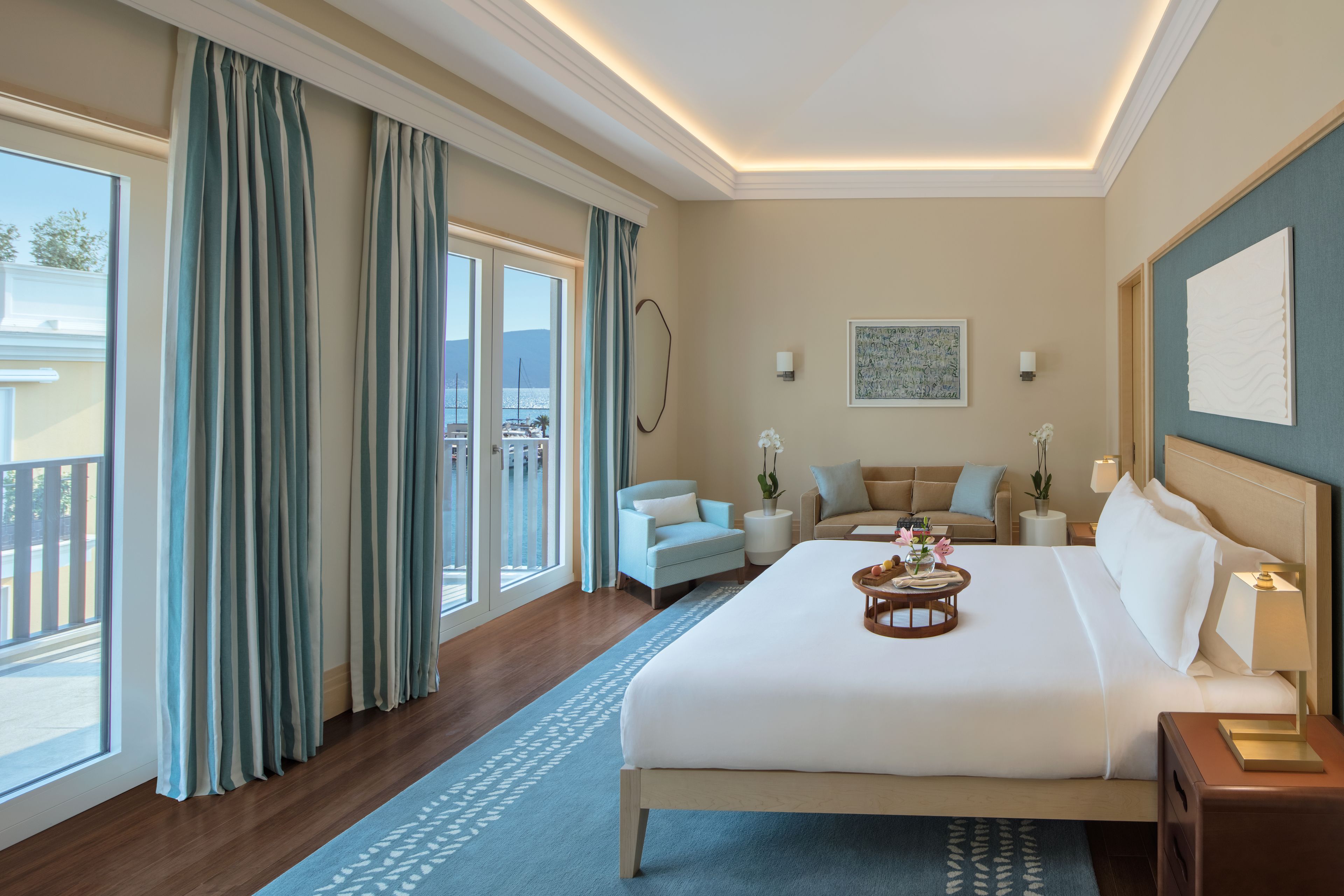 A hotel room with a large bed, a balcony with curtains, a sofa, and a desk with a lamp.
