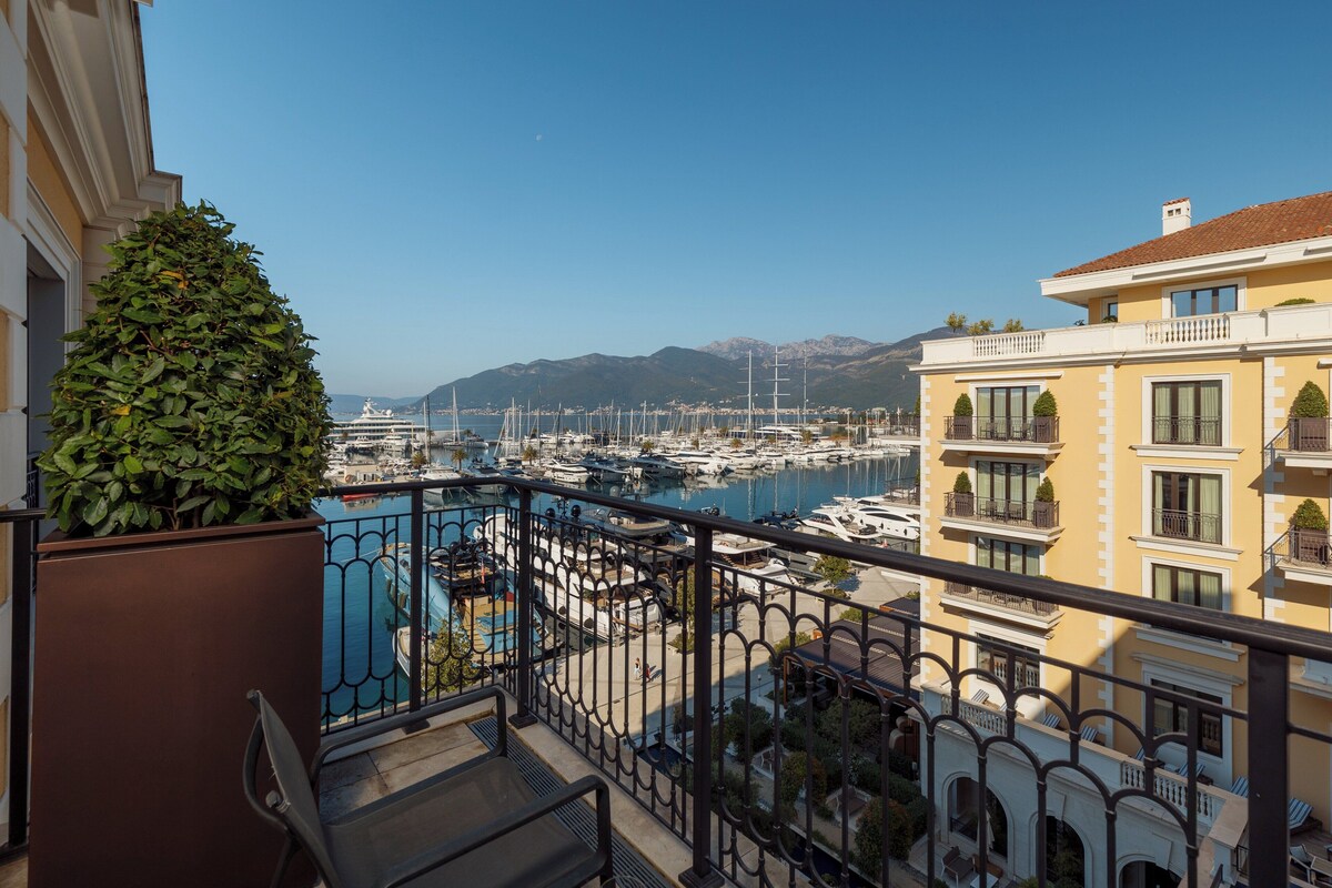 A balcony with a view of a marina, a pool, and a mountain range in the background.