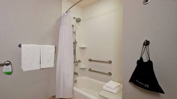 Un baño con bañera, cortina de ducha, toallero con toallas, barra de apoyo y una bolsa negra colgada en la pared.