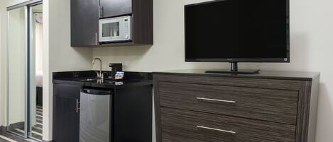 Una kitchenette compacta con microondas, pileta y lavavajillas, un televisor de pantalla plana y un mueble de madera oscura con cajones.