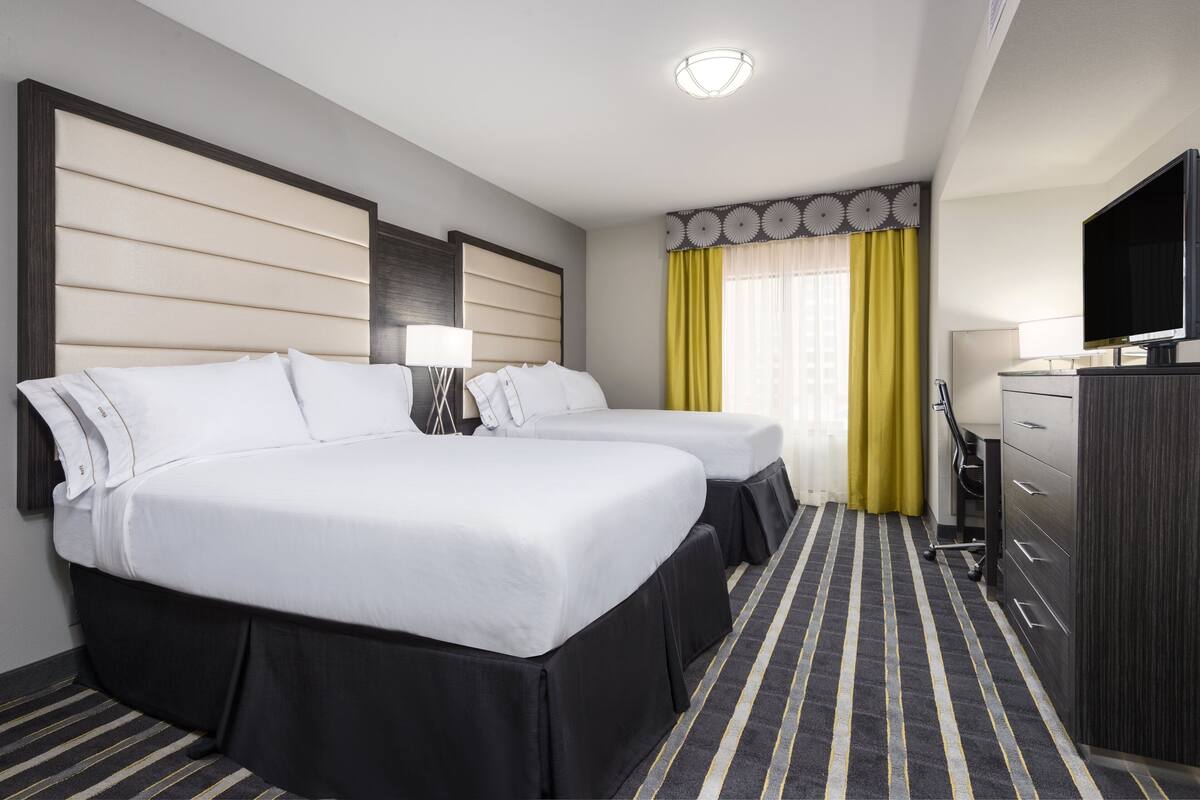 Habitación de hotel con dos camas, un escritorio, televisor y una ventana con cortinas amarillas.