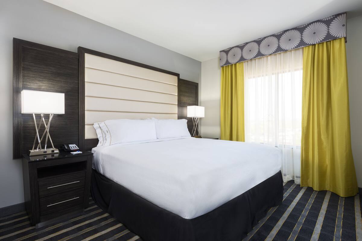 Habitación de hotel con una cama grande, dos mesitas de noche con lámparas, un cabecero de madera oscura y una ventana con cortinas amarillas.