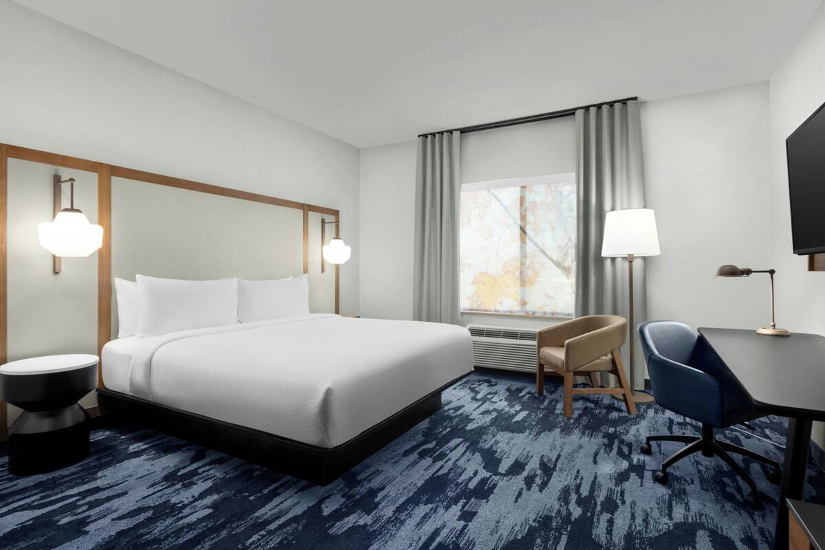 A hotel room with a large bed, a desk, a chair, a TV, and a window with curtains.