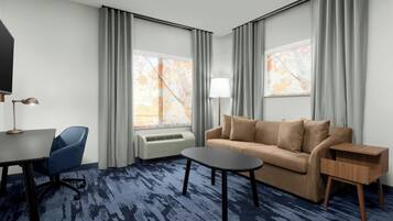 A hotel room with a sofa, a small table, a chair, a TV, and a window with curtains.