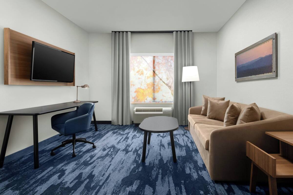 A hotel room with a flat-screen TV, a desk, a chair, a sofa, a coffee table, a lamp, and a painting on the wall.
