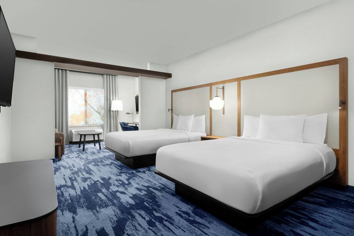 A hotel room with two beds, a desk, a chair, and a large window with curtains.