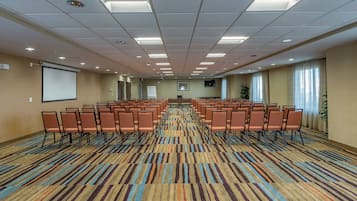 Uma sala de conferência com fileiras de cadeiras, uma tela de projetor, um quadro branco e uma janela grande com cortinas.