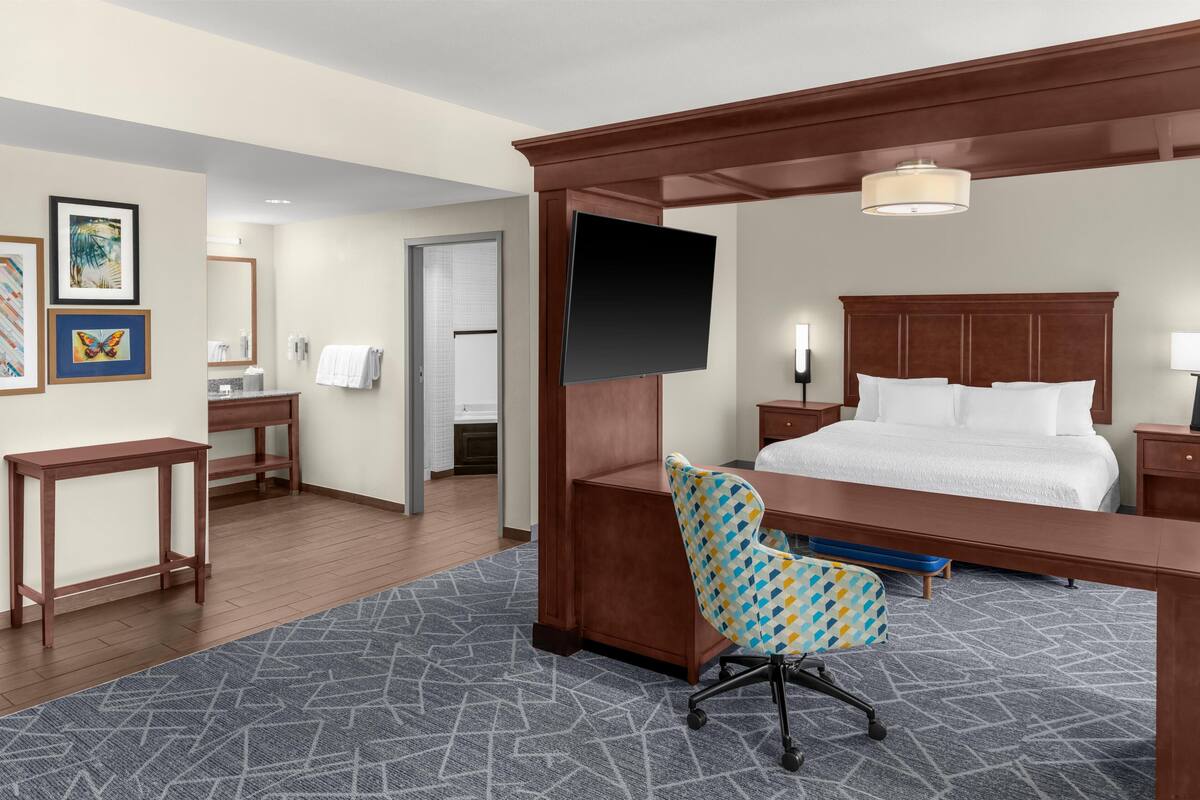 A hotel room with a bed, a desk, a chair, a TV, and a bathroom visible through an open door.