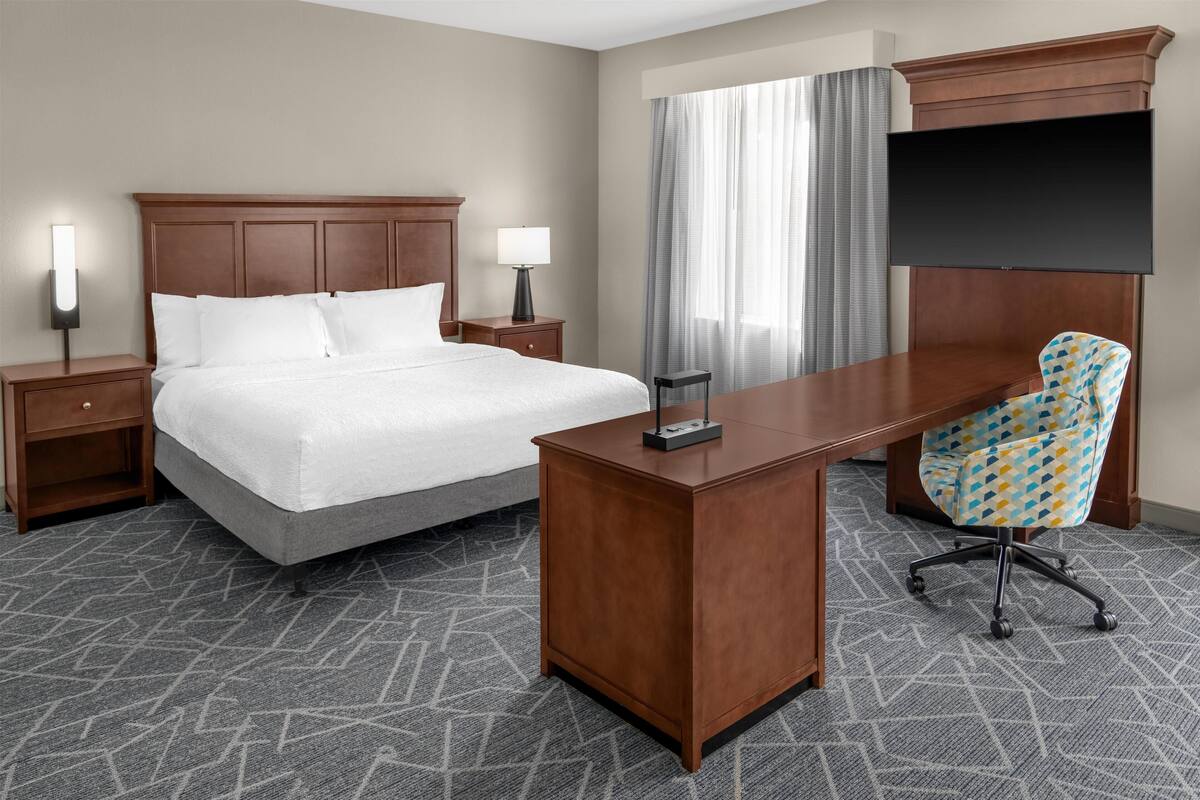 A hotel room with a bed, a desk, a chair, a TV, and a nightstand with a lamp.