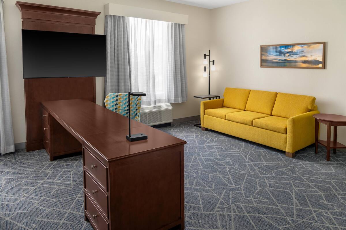 A hotel room with a wooden desk, a yellow sofa, a flat-screen TV, a painting on the wall, and a patterned carpet.