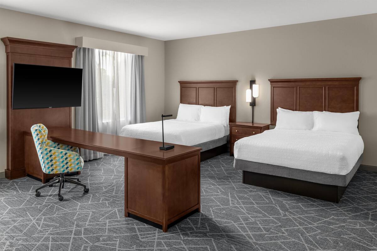 A hotel room with a large desk, two beds, a TV, and a chair.