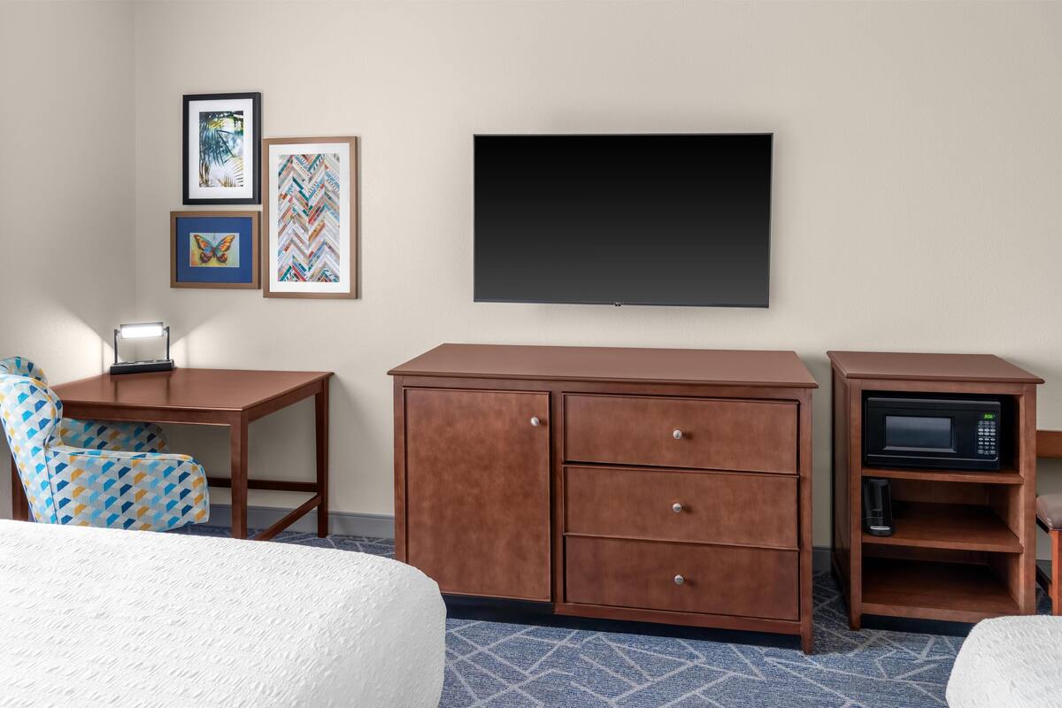 A hotel room with a bed, a desk with a lamp, a dresser, a microwave, and framed artwork on the wall.