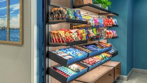 A snack display with various packaged foods and a plant on the shelf. - Hampton Inn & Suites Coconut Creek (Coconut Creek)