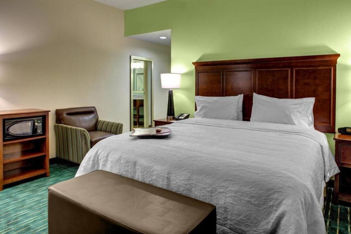 A hotel room with a bed, a chair, a desk with a lamp, and a small table with a tray.