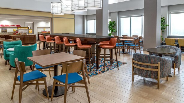 A modern dining area with wooden tables and chairs, a kitchen in the background, and large windows providing natural light. - Hampton Inn & Suites Coconut Creek (Coconut Creek)