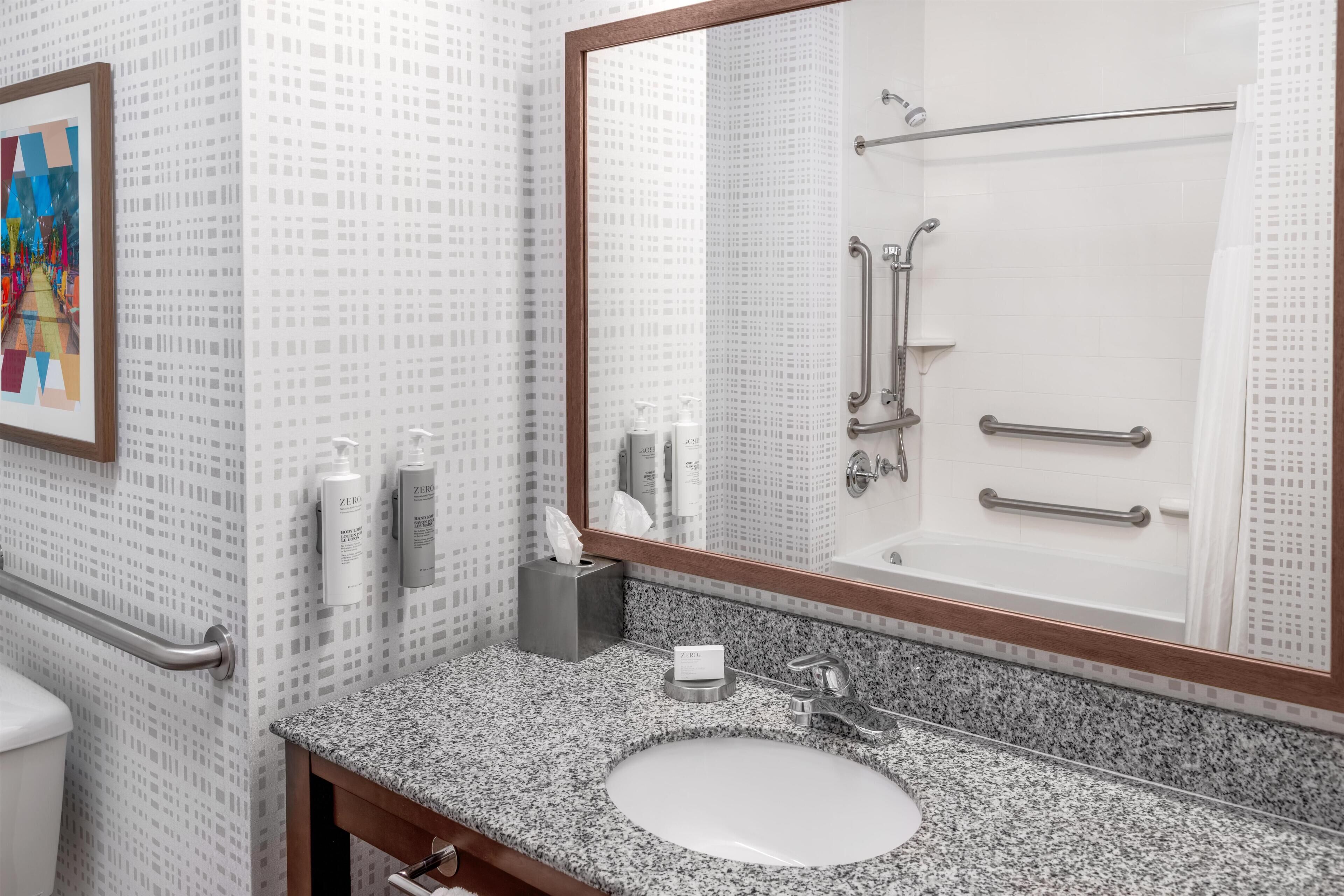 A bathroom with a granite countertop sink, a large mirror, and a shower with grab bars.