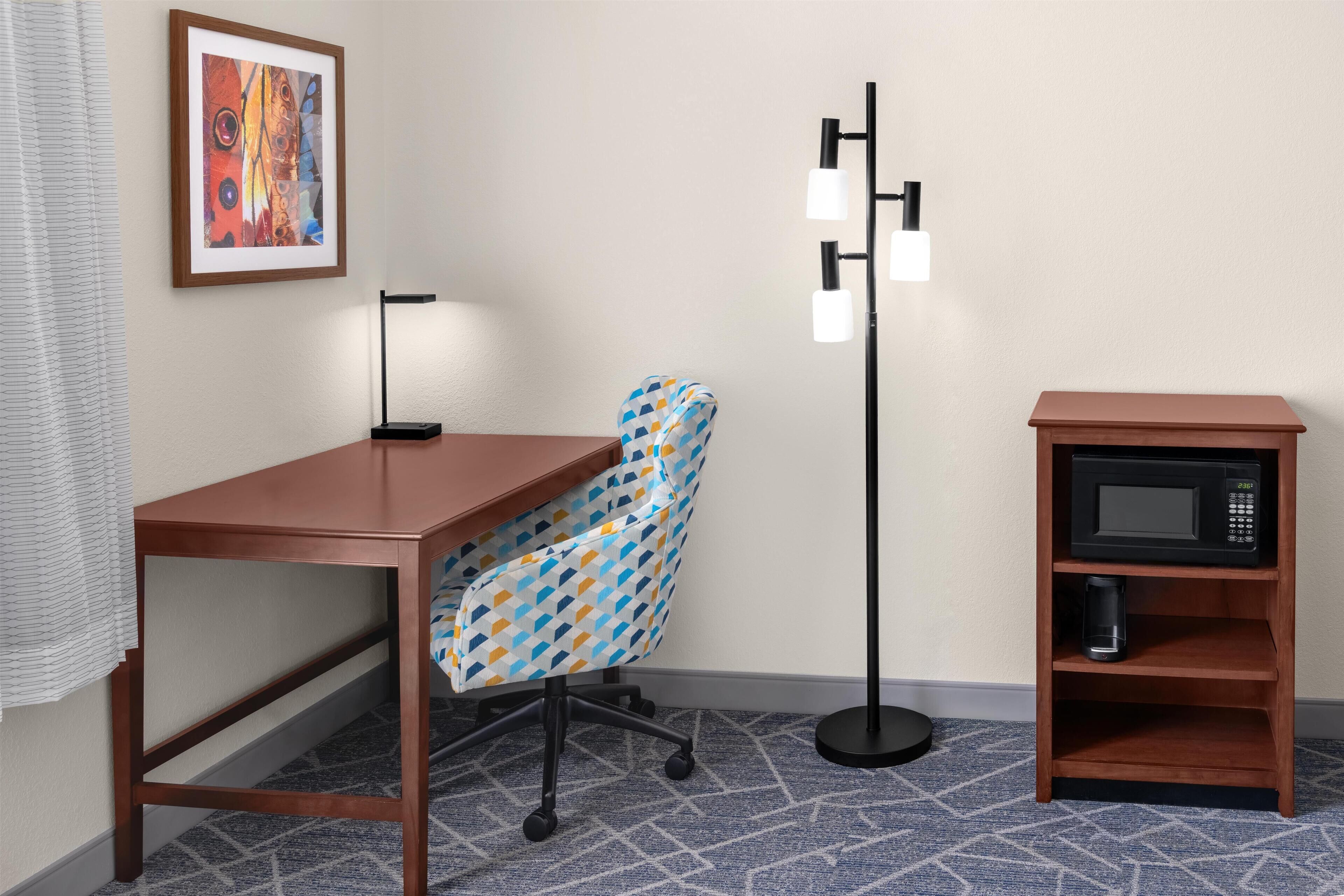 A room with a wooden desk, a patterned office chair, a standing floor lamp, and a microwave shelf.