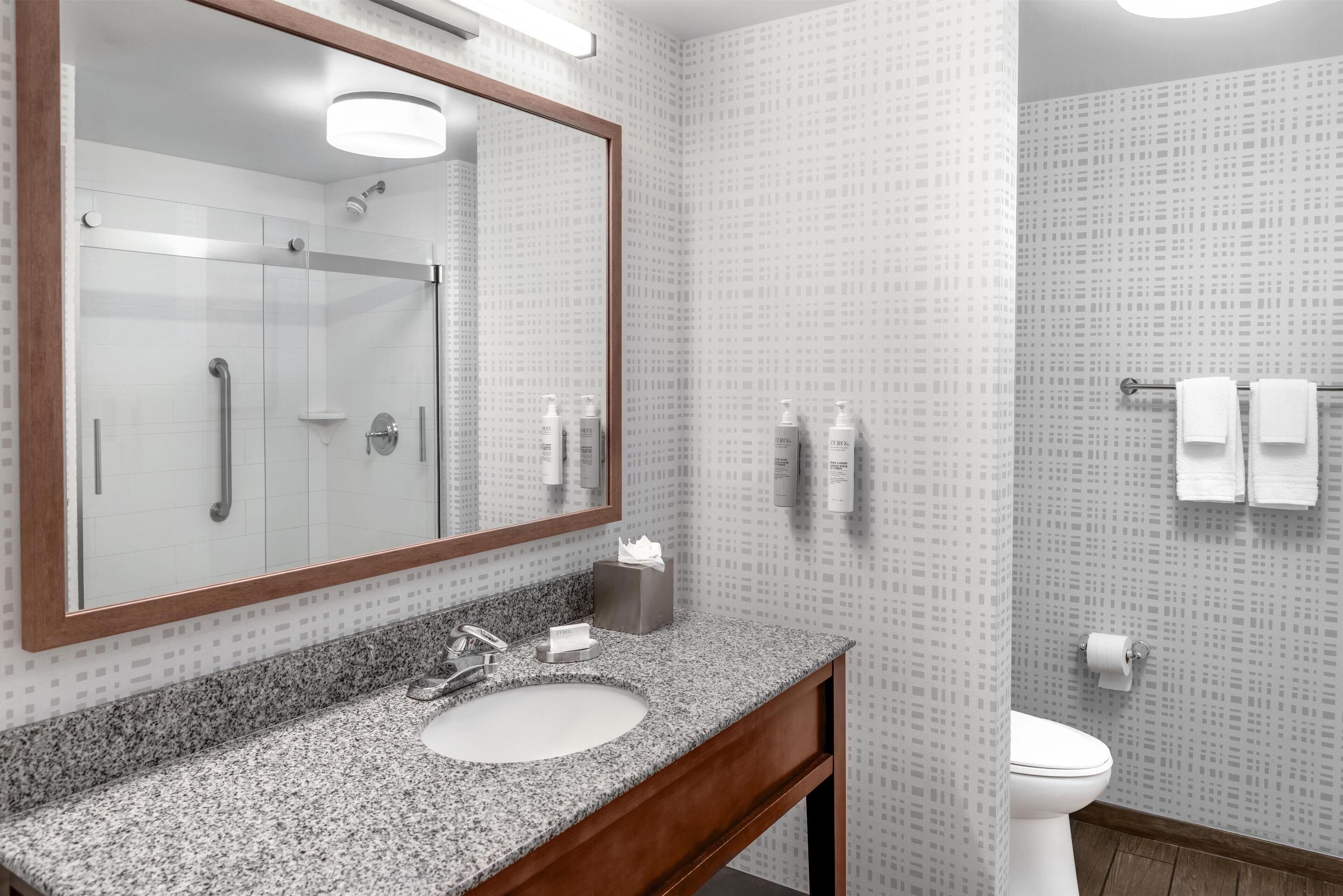 A bathroom with a granite countertop sink, a large mirror, a shower, and a toilet.