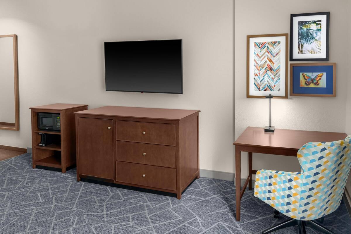 A room with a flat-screen TV, a wooden cabinet, a desk, and a patterned chair.