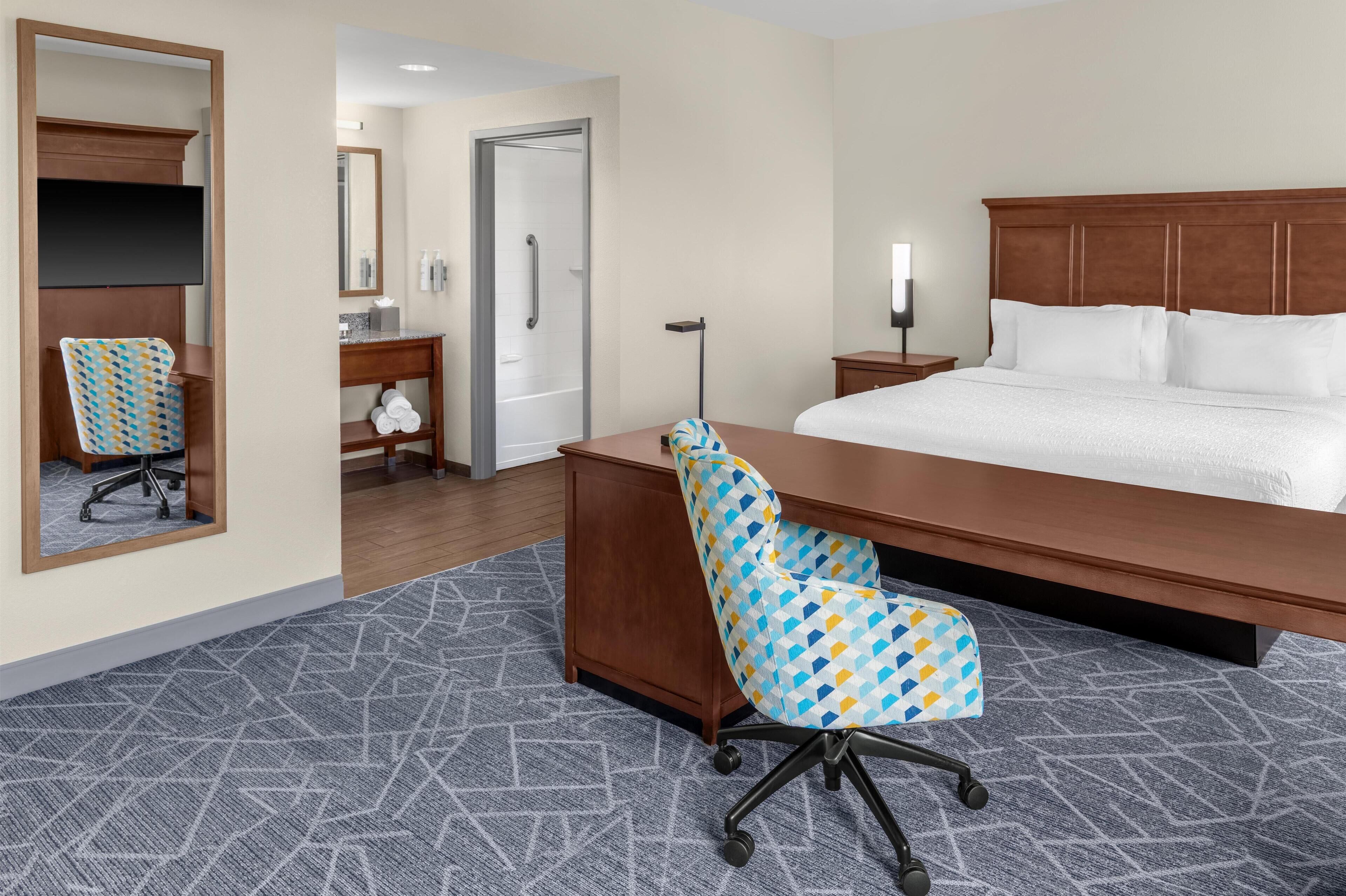 A hotel room with a large bed, a desk with a chair, a mirror, and a bathroom visible through an open door.