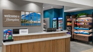 A hotel reception area with a wooden reception desk, a 'Welcome to Hampton' sign, a 'Hilton Honors' sign, a refrigerator, and a snack display. - Hampton Inn & Suites Coconut Creek (Coconut Creek)