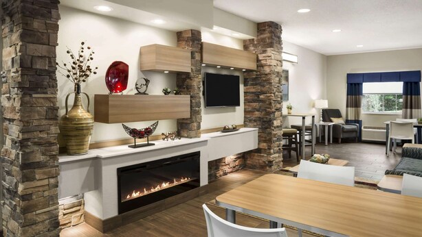 A modern living room with a stone fireplace, a flat-screen TV, and a dining area with a wooden table and chairs. - Microtel Inn & Suites By Wyndham Lynchburg (Lynchburg)